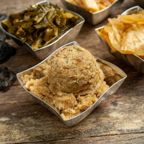 A close-up of cornbread dressing in a silver bowl, with collard greens and cabbage in the background.